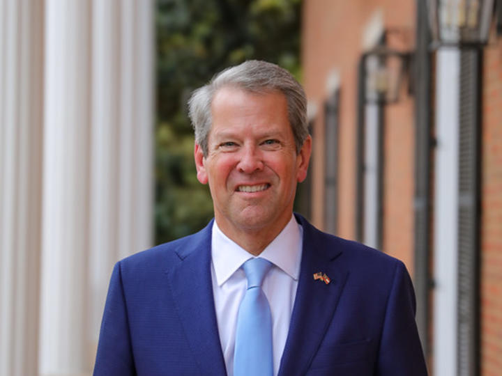 Governor Brian Kemp wears a suit a tie smiling in front of a large building